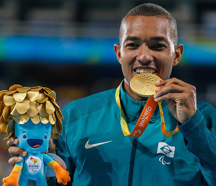 Descrição da imagem: Ricardo morde medalha e exibe o mascote Tom com cabelos dourados (Foto: infoesporte)